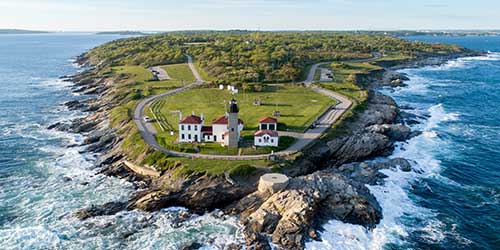 Aerial view of Beavertail Lighthouse surrounded by rocky coastline and ocean waves in Rhode Island.