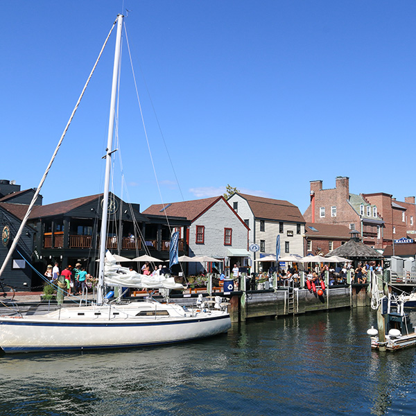Bowen’s Wharf in Newport, Rhode Island with a sailboat docked along the harbor and waterfront restaurants and shops bustling with visitors on a sunny day.