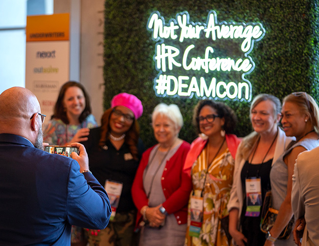Group of women posing for a photo in front of a sign that reads Not Your Average HR Conference #DEAMcon
